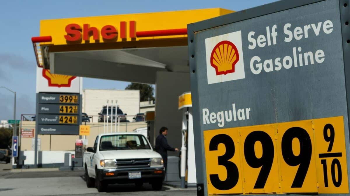 A wide shot of a Shell gas station under a clear blue sky, featuring a large grey sign on the right with the Shell logo and "Self Serve Gasoline," showing "Regular" fuel priced at $3.99 and 9/10 cents per gallon in black numbers on yellow panels. In the background, a yellow and red Shell canopy covers fuel pumps. A white pickup truck is visible at one of the pumps, with a person standing nearby. A tall sign under the canopy also shows gas prices: Regular at $3.99 and 9/10, Plus at $4.12 and 9/10, and V-Power at $4.24 and 9/10.