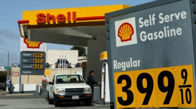 A wide shot of a Shell gas station under a clear blue sky, featuring a large grey sign on the right with the Shell logo and "Self Serve Gasoline," showing "Regular" fuel priced at $3.99 and 9/10 cents per gallon in black numbers on yellow panels. In the background, a yellow and red Shell canopy covers fuel pumps. A white pickup truck is visible at one of the pumps, with a person standing nearby. A tall sign under the canopy also shows gas prices: Regular at $3.99 and 9/10, Plus at $4.12 and 9/10, and V-Power at $4.24 and 9/10.