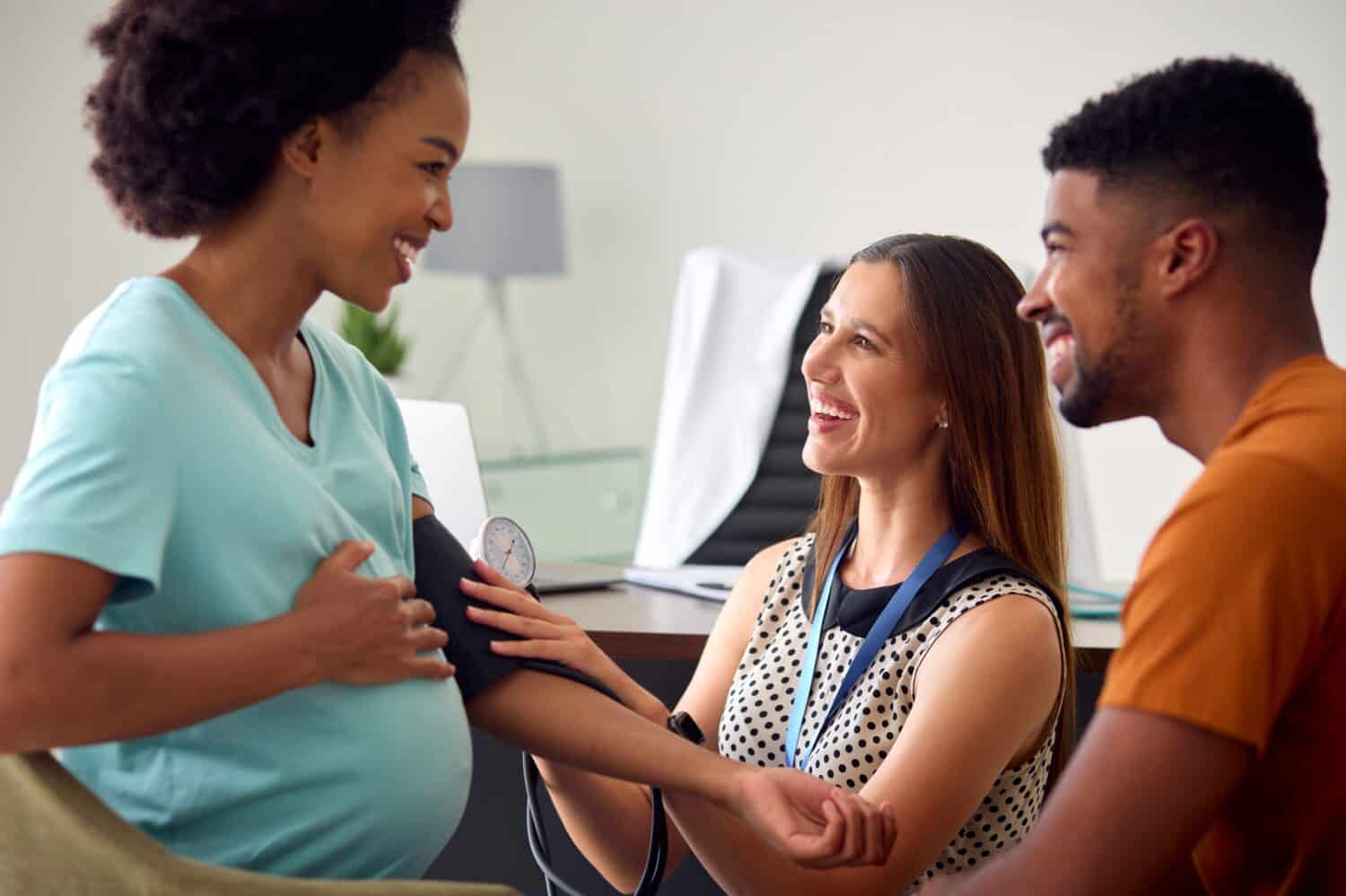 Pregnant Couple At Appointment With Female Doctor Or GP Taking Blood Pressure In Office