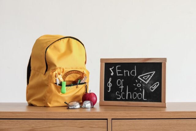 Chalkboard with text END OF SCHOOL, apple and backpack on table near light wall