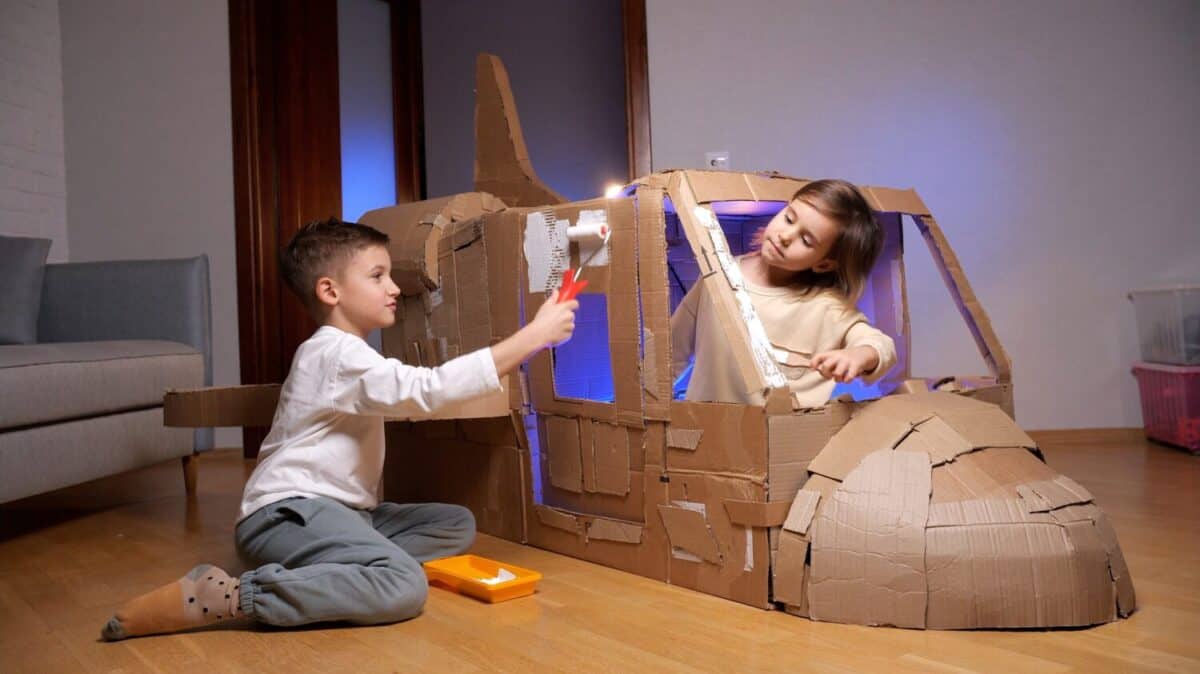 Two young children painting a cardboard spaceship they built together. They are adding a fresh coat of white paint to their cardboard creation