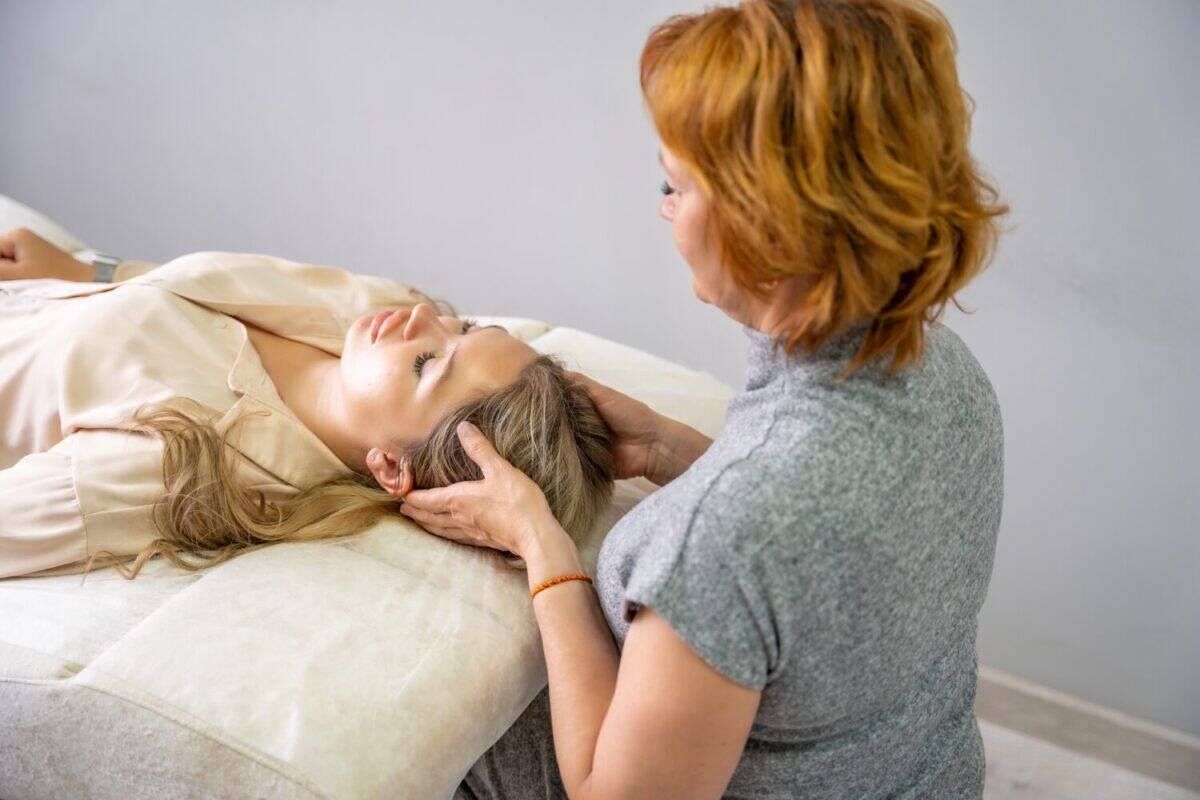 Female practitioner performs facial lifting technique on a young woman lying on a couch. Concept of energy healing, face therapy and alternative wellness practice from Access Bars. High quality photo