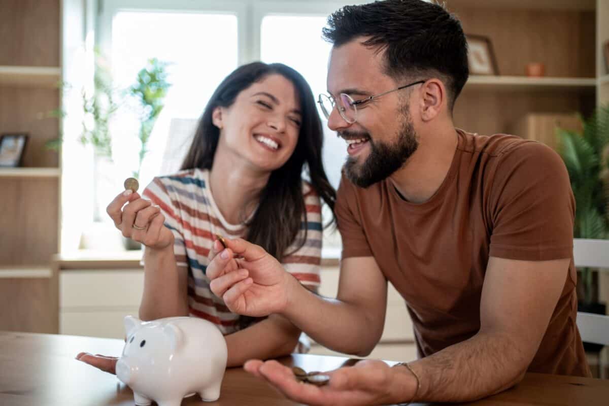 Young smiling couple putting coins into piggy bank, saving money for future, managing home budget, making financial plans together, successful investment strategy, happy family life