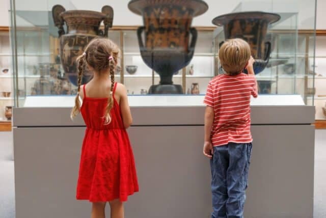 In a museum, a young boy and girl stand by a case featuring massive antique pottery, their attention drawn to the detailed craftsmanship