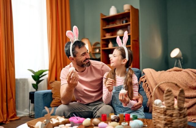 A family tradition on the eve of Easter. A father and his daughter, dressed in pink outfits and wearing bunny ears, are decorating eggs in a bright and colorful living room.