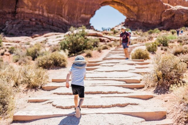 A young child running up the stair pathway to the Turret Arch at Arches National Park in Moab, Utah on a hot summer day with his dad blurry and in the background.