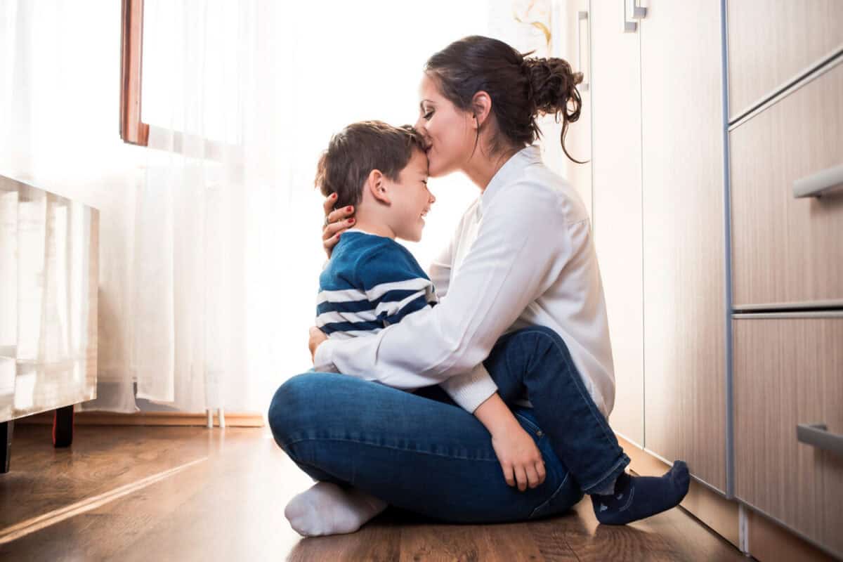 Beautiful young mom and her son, sitting on the floor hugging each other tightly. Mom is kissing her kid in the forehead. Mother kid relationship