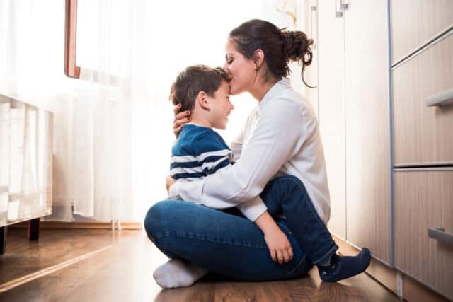 Beautiful young mom and her son, sitting on the floor hugging each other tightly. Mom is kissing her kid in the forehead. Mother kid relationship