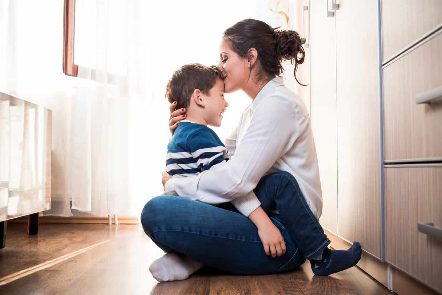 Beautiful young mom and her son, sitting on the floor hugging each other tightly. Mom is kissing her kid in the forehead. Mother kid relationship