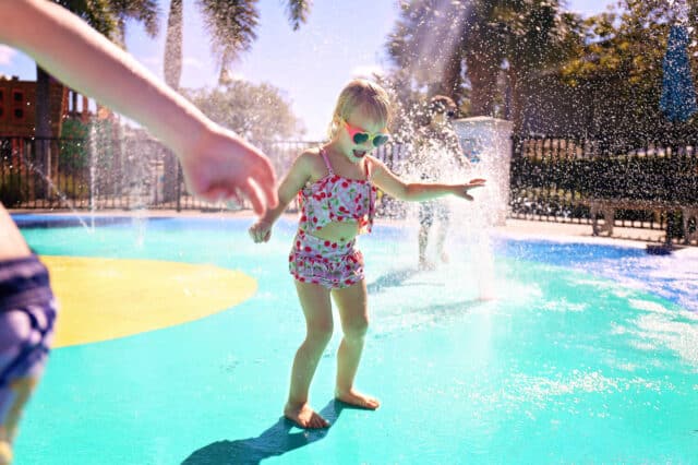 Little kids are playing outside in the water fountains at an outdoor splash pad park on a summer day.