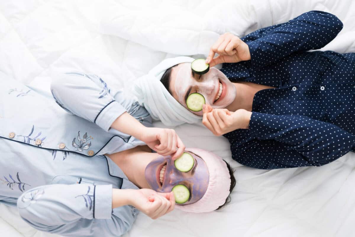 Top view of two excited women friends in bed putting cucumber slices in their eyes to finish a relaxing skin care routine during a fun sleepover