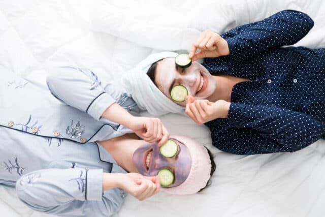 Top view of two excited women friends in bed putting cucumber slices in their eyes to finish a relaxing skin care routine during a fun sleepover