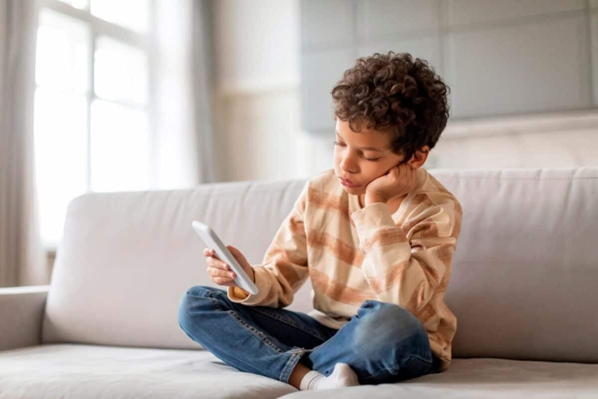 Upset Little Black Boy Using Mobile Phone While Relaxing On Couch At Home, Bored Preteen Male Child Playing Games Or Browsing Internet On Smartphone, Sad Kid Checking New App, Copy Space