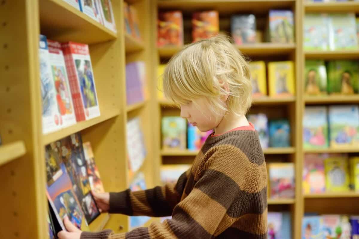A preteen boy leafing through a book while standing at the bookshelfs in a school library or bookstore. Smart kid reading comics or adventure book