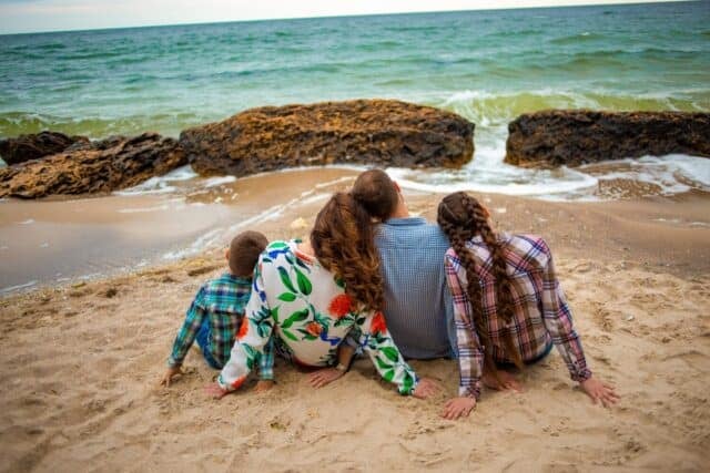 A family of four sitting on the beach with their backs to the sea, gazing out at the ocean together, capturing a moment of togetherness and serenity