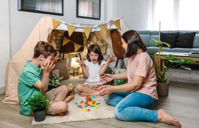 Happy family laughing and shouting as a tower of wooden stacking piece game falls. Mother and sons having fun while playing together in front of homemade teepee on living room. Staycation concept.