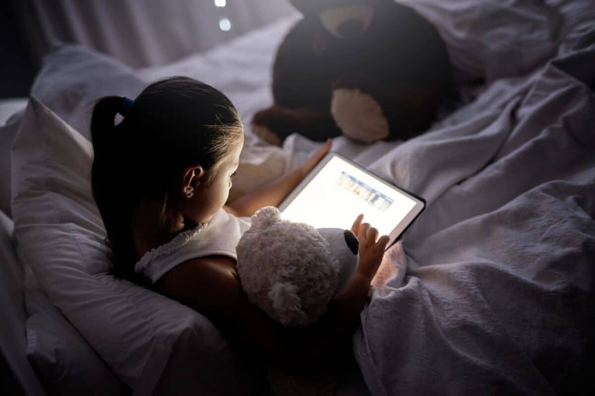 Tablet, reading and girl in bed at night with ebook, story and youth game with cartoon and language app. Tech, teddy bear and young kid with scroll, relax and evening screen time at home on internet