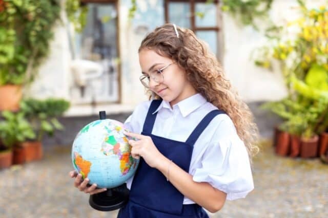 Smiling young girl in glasses and in a school uniform holding globe for geography lessons. Back to school concept. Portrait of cheerful girl with curly hair and in casual clothes holding globe.
