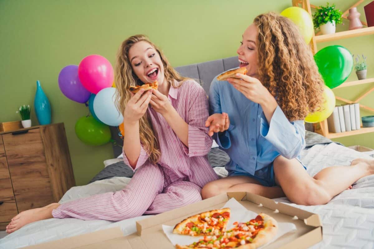 Full length photo of two pretty young females sit bed eating pizza dressed enjoy wake up modern cozy day light bedroom home interior