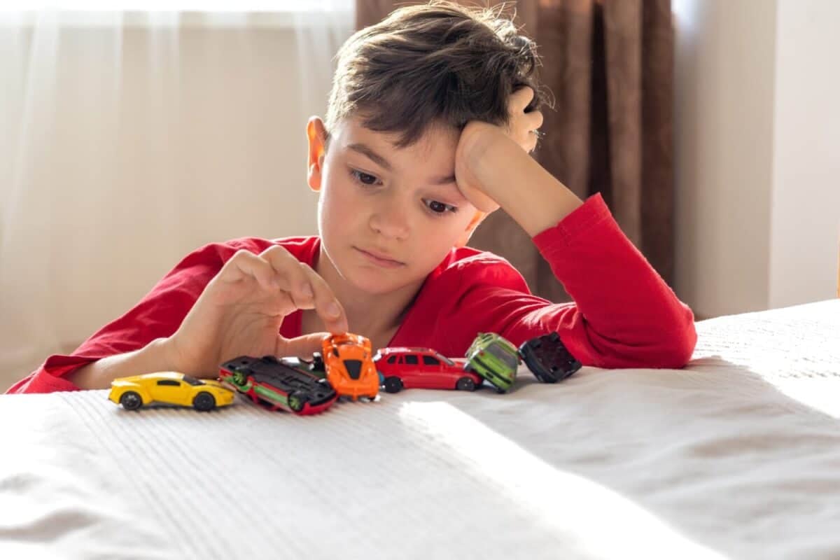 A bored boy in a red shirt plays with toy cars on his bed, appearing uninterested. This scene suggests ADHD, autism, or emotional challenges, reflecting child development and mental health issues.