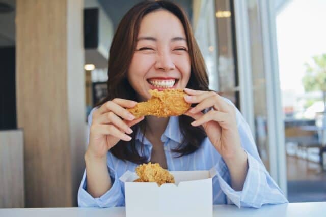 A joyful young Asian woman enjoying a piece of fried chicken at a modern restaurant. She laughs and holding the crispy food.