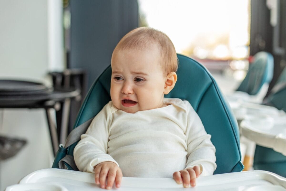 Distressed baby sitting in a high chair, appearing uncomfortable or hungry, during mealtime in a brightly lit indoor space.