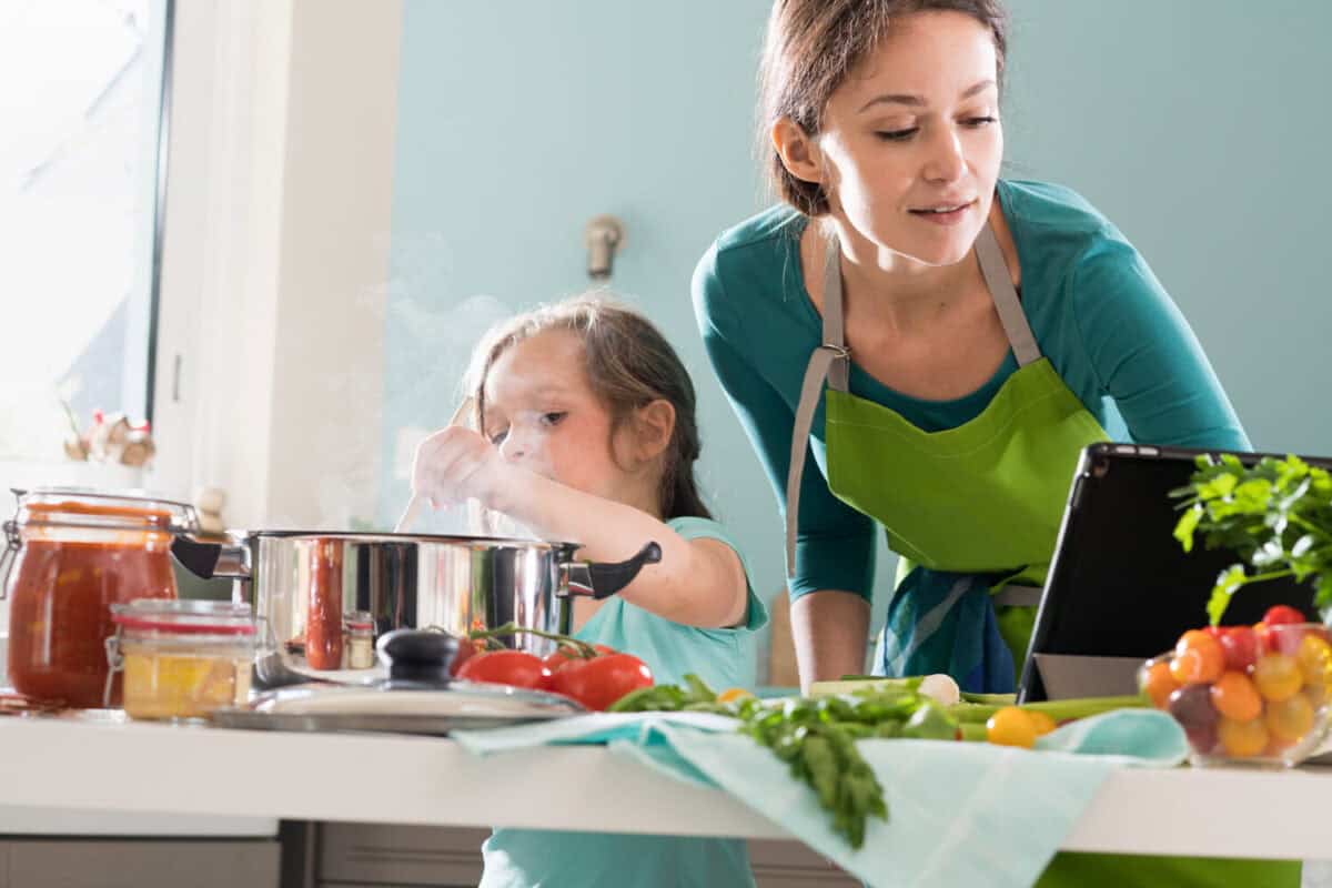 A mother and her daughter using a digital tablet to cook a recipe in the kitchen. The little girl stirs the contents of a saucepan with a spoon, there is steam above the pan
