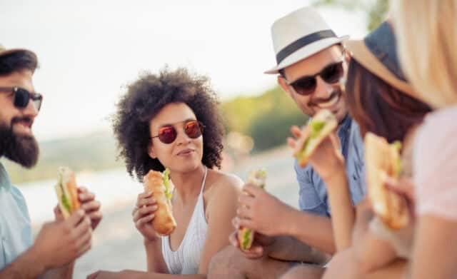 Group of friends sitting on the beach and eating sandwiches during picnic.