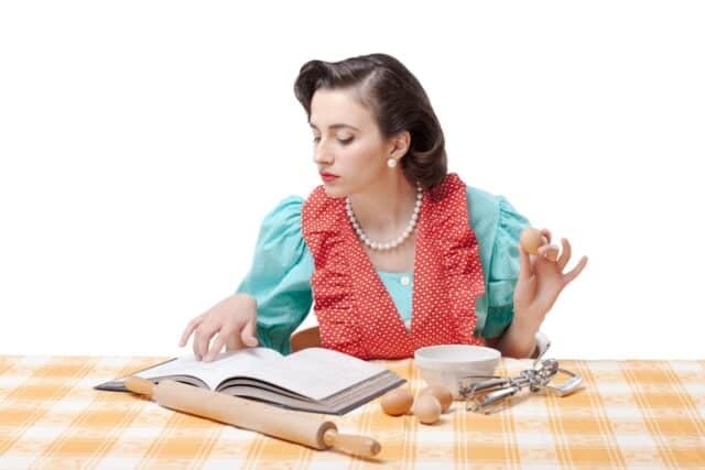 Beautiful vintage style housewife preparing a dessert at home, she is checking the recipe in a cookbook and mixing ingredients in a bowl
