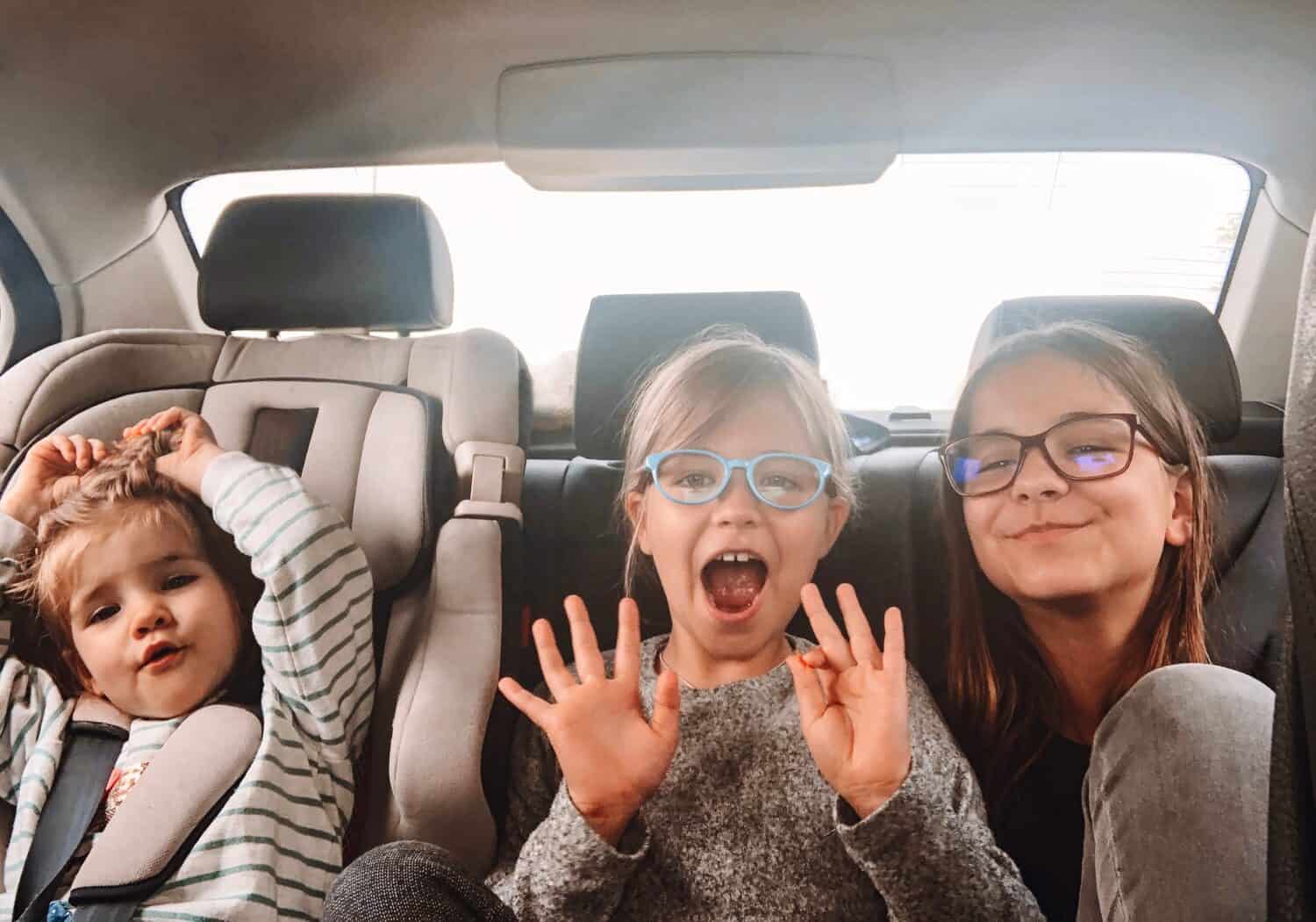 Three cute sisters of different ages are riding in the back seat of a car. Girls scream, indulge and laugh. Road trip with three children. Kids having fun in the automobile. Free space for the text.