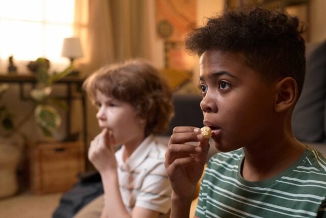 Two diverse children sitting on floor eating popcorn while watching TV in cozy old-fashioned living room with focused facial expression