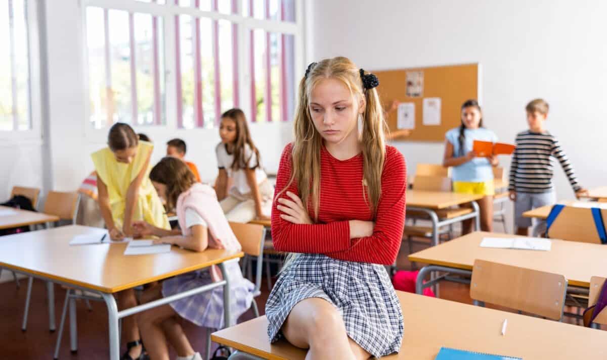 Portrait of sad girl sitting alone on desk during recess in school.