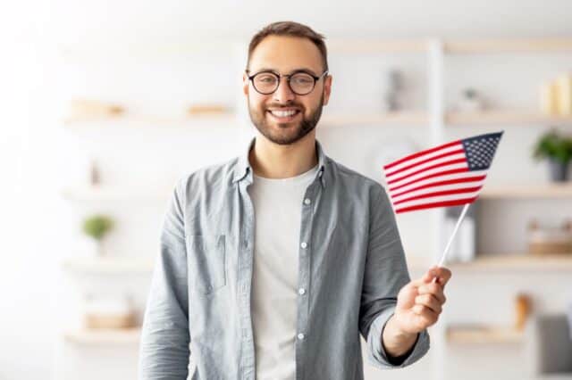 Handsome young guy holding American flag and smiling at camera indoors. Cheerful millennial man recommending English language school, emigrating to United States. Abroad education and student exchange