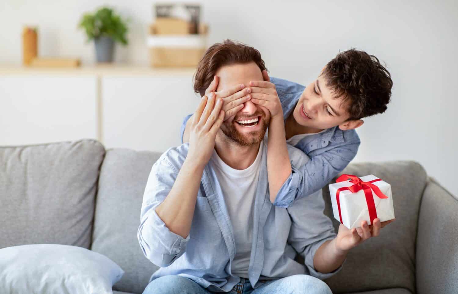 Happy father's day. Son congratulating his daddy, covering his eyes from behind, greeting and giving present at home. Family holiday celebration, surprise present for dad