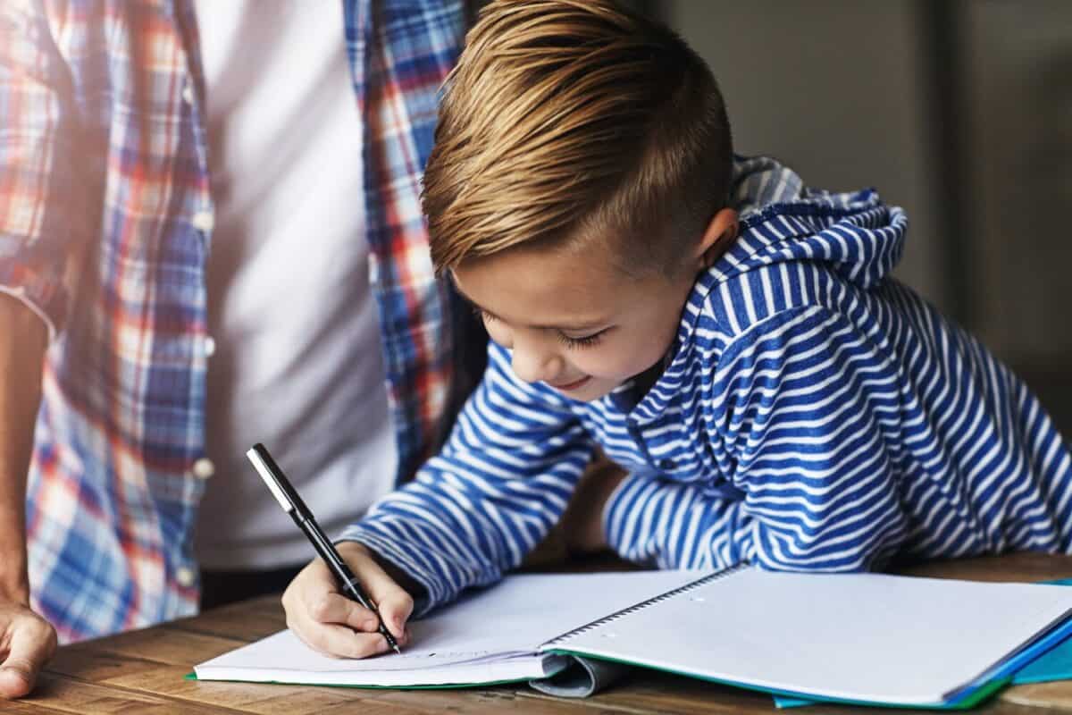 Writing, child or parent with homework help in home for study support, literacy lesson or learning. Education, family or kid with book at table for handwriting practice, fine motor skills or guidance