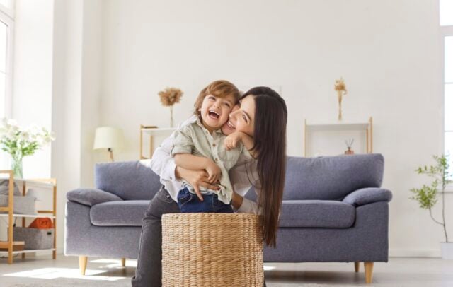 Happy mother and child play at home, bonding. A playful toddler kid laughs in a wicker basket as woman parent smiles nearby, sharing love and happiness. Warm family leisure and joy together.