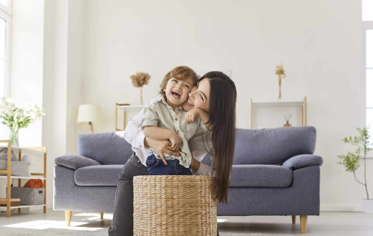 Happy mother and child play at home, bonding. A playful toddler kid laughs in a wicker basket as woman parent smiles nearby, sharing love and happiness. Warm family leisure and joy together.