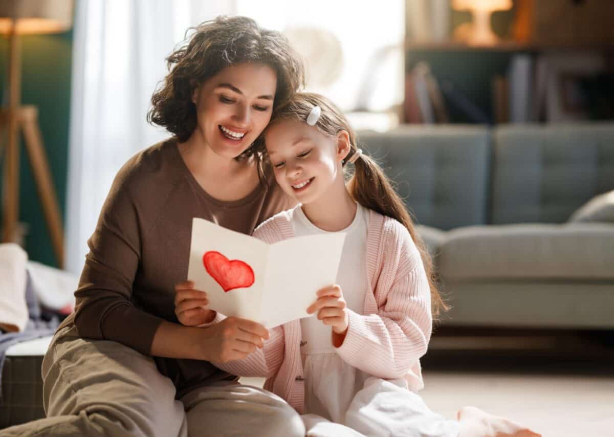 Joyful mother and daughter share a heartwarming moment at home, reading a handmade card together. The cozy setting, soft lighting, and genuine smiles capture the essence of family love, bonding, and special occasions. Perfect for concepts of Mother's Day, family relationships, affection, and cherished childhood memories.