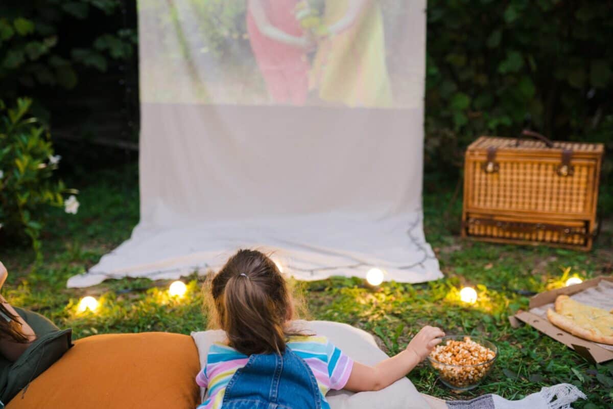 Family summer outdoor movie night. Girl lying on blanket, eating homemade popcorn and watching film on DIY screen with from projector. Summer outdoor weekend activities with kids. Open air cinema