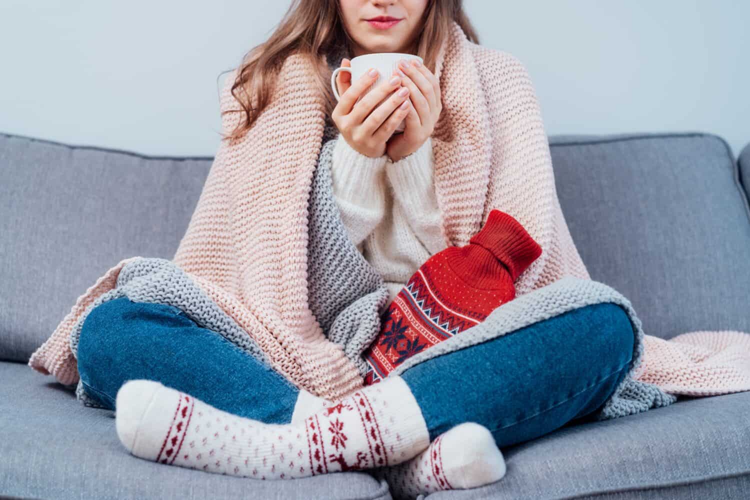 Woman freezes in wintertime. Young girl wearing warm woolen socks and wrapped into two blankets, holding a cup of hot drink and heating pad while sitting on sofa at home. Keep warm. Selective focus
