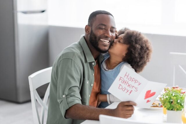 curly african american girl kissing cheek of happy father with greeting card on fathers day