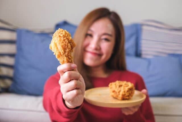 Portrait image of a young woman holding and eating fried chicken at home