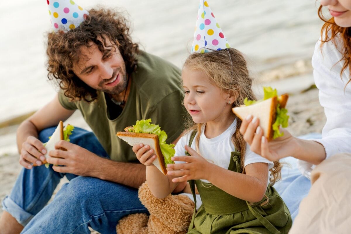 Close up shot of family with party hats eating sandwiches on the beach. Family enjoying daughter's birthday with snacks outdoors. Healthy eating, vegetarian meal. Food festival