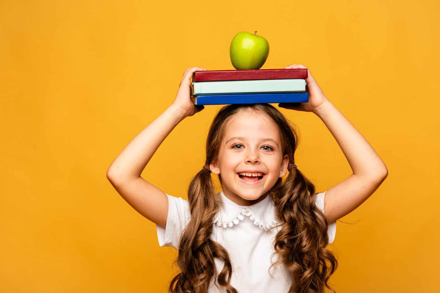 Funny happy Preschool Child girl with Book and green apple on Head and Bag on Yellow Background, Copy Space. Happy Smiling Kid Go Back to School, Kindergarten. Success, Motivation. School lunch time