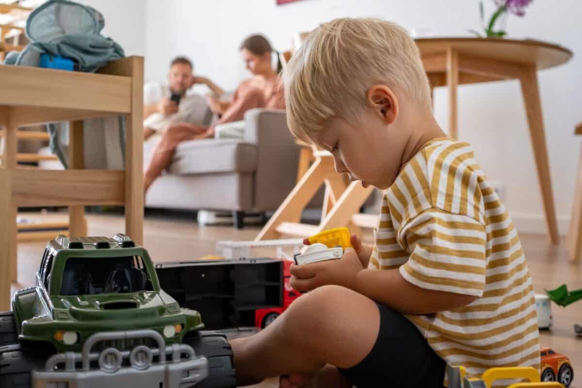 A young child sits on the floor, deeply focused on playing with various toy trucks. Parents relax on the sofa in the blurred background. Scene captures independent, imaginative playtime at home.