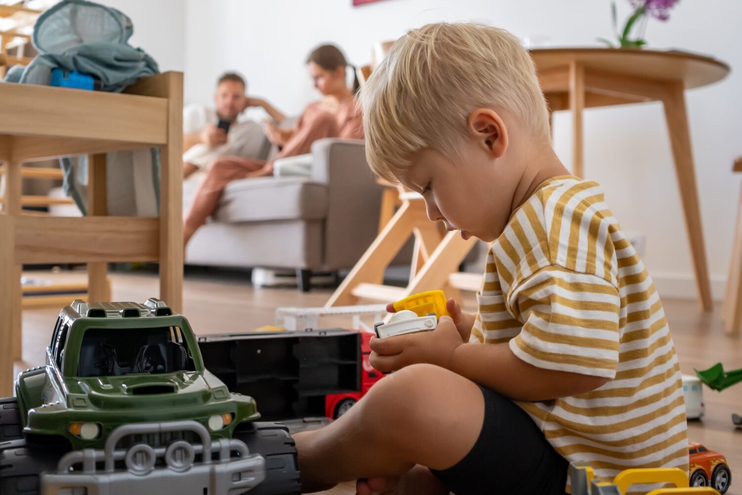 A young child sits on the floor, deeply focused on playing with various toy trucks. Parents relax on the sofa in the blurred background. Scene captures independent, imaginative playtime at home.