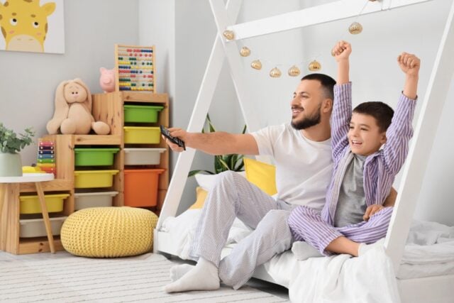 Happy father and his son in pajamas watching TV on bed at home
