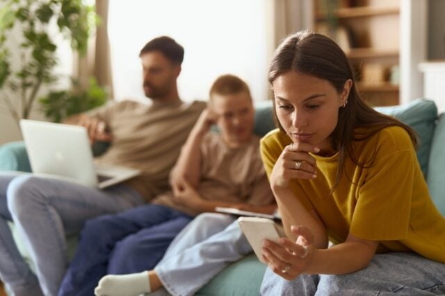 Mother text messaging on smart phone while relaxing next to her kids and husband on sofa in the living room.