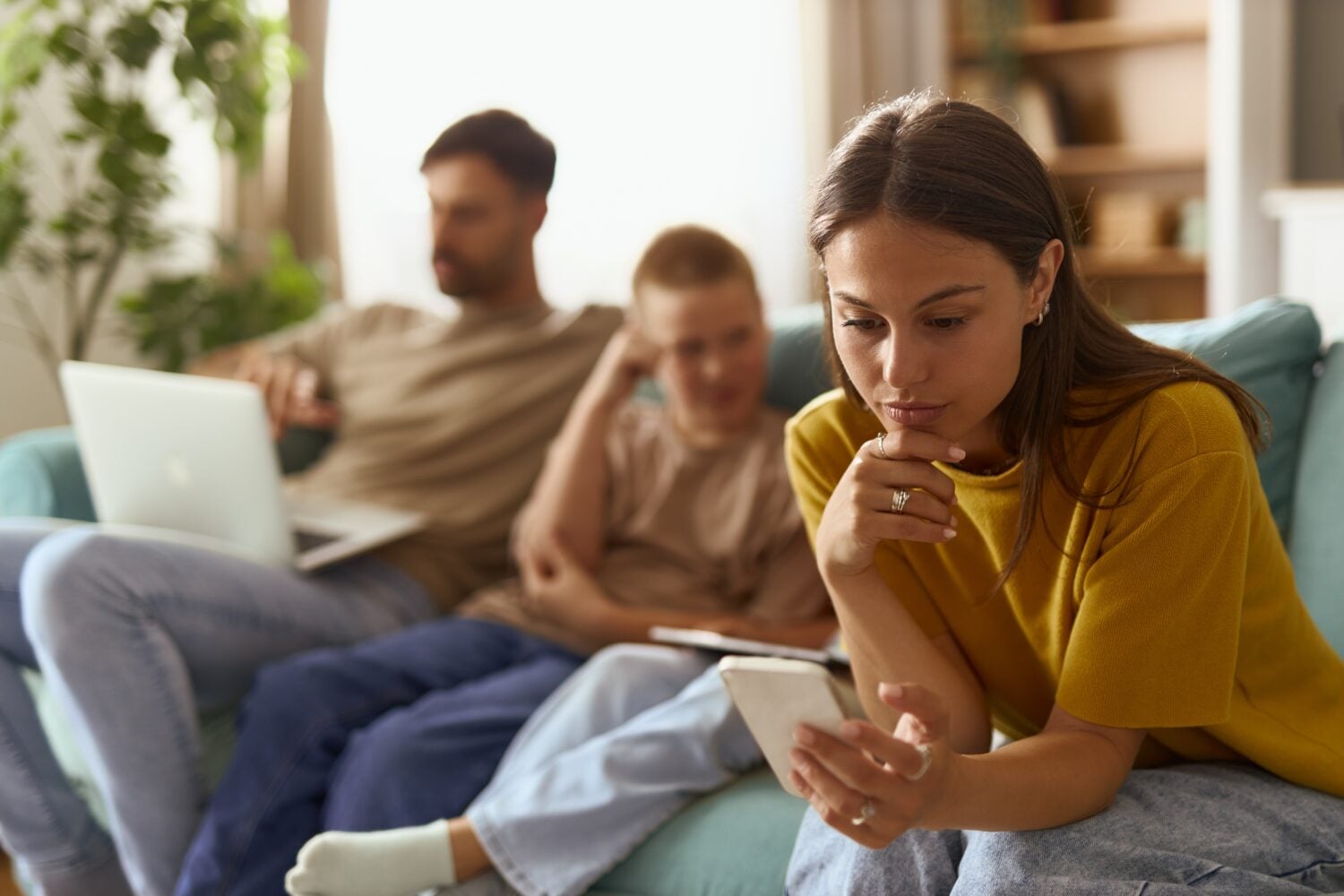 Mother text messaging on smart phone while relaxing next to her kids and husband on sofa in the living room.