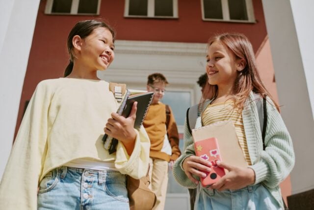 Two preteen girls, one Asian and one Caucasian, smiling and talking while holding notebooks and walking down steps outside school building, boy with backpack following behind